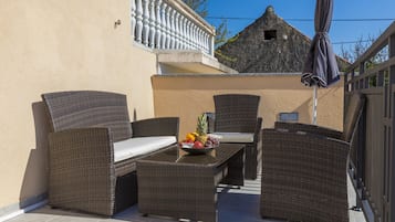 Um pátio externo com móveis de vime, uma mesa com frutas e vista para um edifício ao fundo.