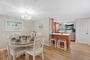 A dining area with a round table, white chairs, and a kitchen in the background. - Folly Field Villa | Walk 2 Beach+Pool | Bikes, BBQ (Hilton Head Island)