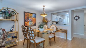 A dining area with a glass table, wooden chairs, a hanging light fixture, a painting, and a kitchen in the background. - Renovated Forest Beach Villa | Walk to Beach & Coligny Plaza (Hilton Head Island)