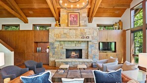 A spacious living room with a stone fireplace, wooden ceiling beams, and large windows.