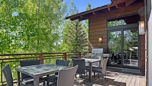 An outdoor patio with a wooden deck, glass table, wicker chairs, and a sliding glass door leading to an indoor area with a grill.