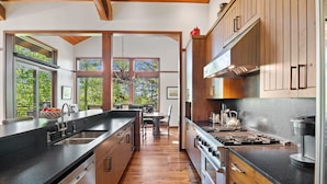 A modern kitchen with wooden cabinets, a black countertop, stainless steel appliances, and large windows offering a view of greenery.