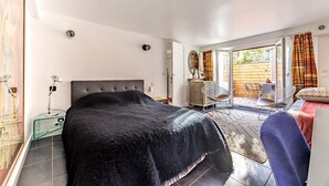 A bedroom with a large bed, a glass side table, a desk, and a chair. There is a sliding glass door leading to an outdoor area with a wooden deck and chairs. - Comfortable studio in a quiet location 10 minutes from the RER station (Joinville le pont)