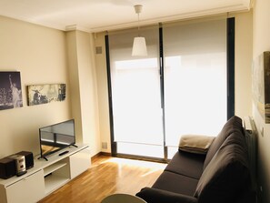Ein modernes Wohnzimmer mit Flachbildfernseher, einer Couch, einem Couchtisch und einem großen Fenster mit Jalousien.