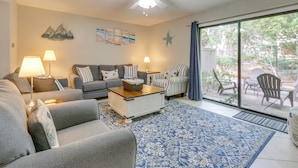 Ein Wohnzimmer mit grauem Sofa, Couchtisch und gestreiften Sesseln. Es gibt einen Deckenventilator, ein großes Fenster mit Blick ins Freie und einen blau gemusterten Teppich.