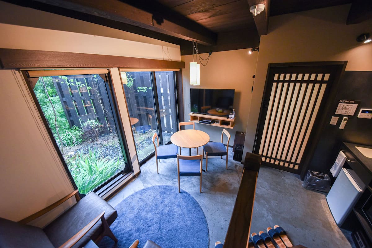 A room with a large sliding glass door leading to a garden, a round table with chairs, a small TV, and a refrigerator.