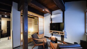 Een traditionele Japanse kamer met tatami vloeren, een lage houten tafel, twee houten stoelen, een aan de muur bevestigde televisie en een schuifdeur naar een andere kamer.
