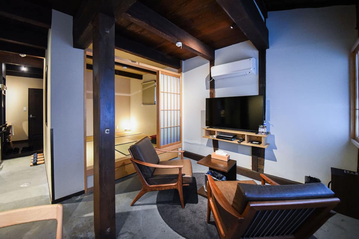 A traditional Japanese-style room with tatami flooring, a low wooden table, two wooden chairs, a television mounted on the wall, and a sliding door leading to another room.