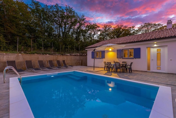 Una piscina con sillones de descanso, una mesa con sillas y un edificio de tejado con baldosas.