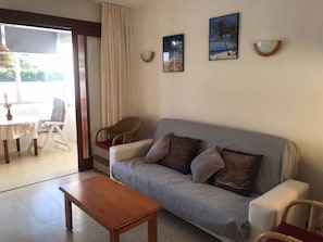 A living room with a sofa, a wooden table, and a wicker chair. There are two wall-mounted light fixtures and a window with curtains.