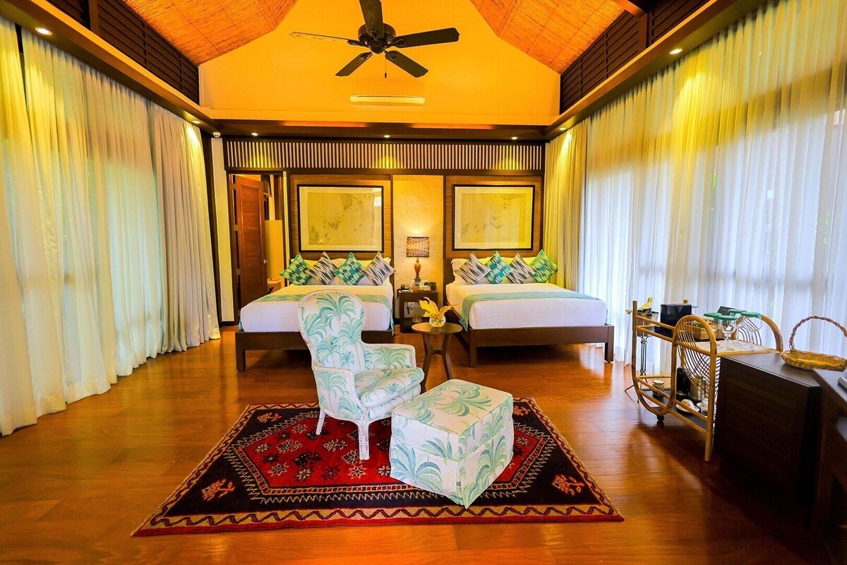 A hotel room with two beds, a ceiling fan, a large window with curtains, a small table, and a chair.
