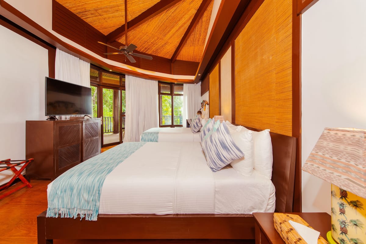 A bedroom with a large bed, a television, a ceiling fan, and a window with curtains.