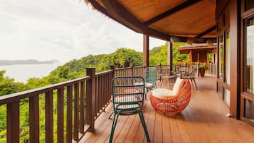 Terraza de madera con sillas de mimbre y una zona de estar acolchada con vistas al agua y la vegetación.