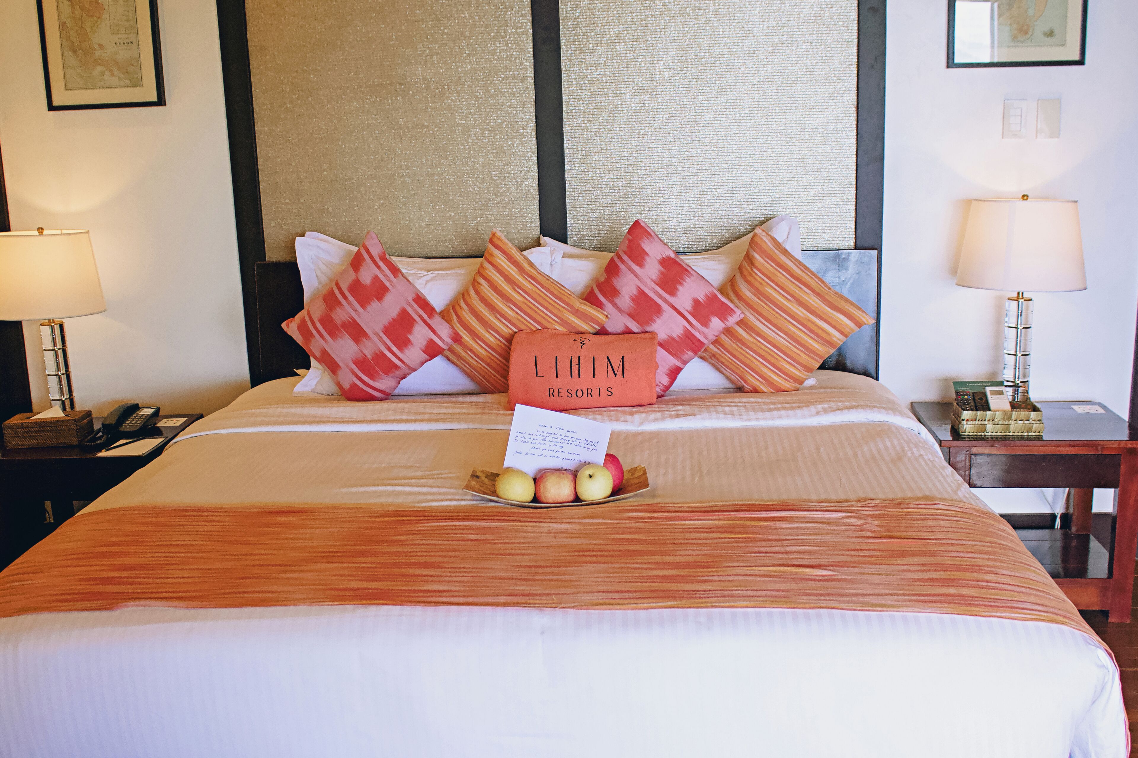 Una cama bien hecha con un cartel que dice 'LIHIM RESORTS', una mesita de noche con una lámpara, y un frutero con manzanas sobre la cama.