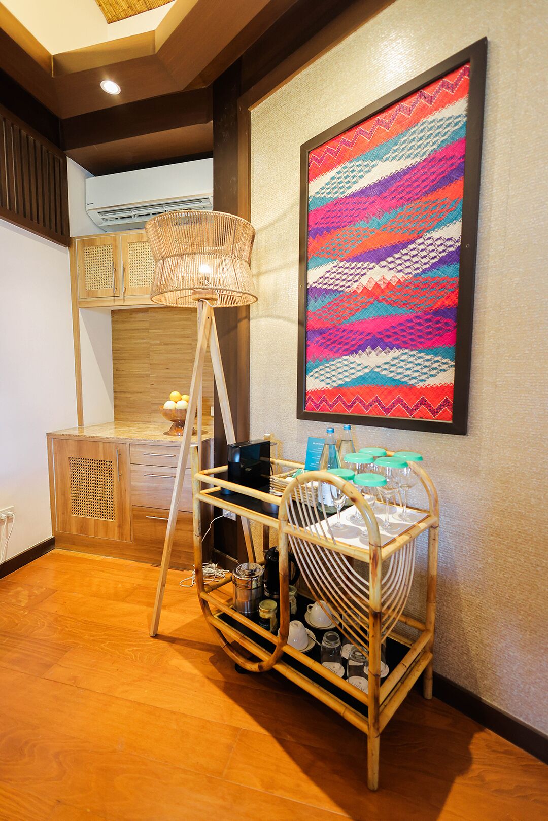 Una habitación con un armario de madera, una lámpara de pie tejida, un carrito de comedor con cristalería y un tapiz de colores.