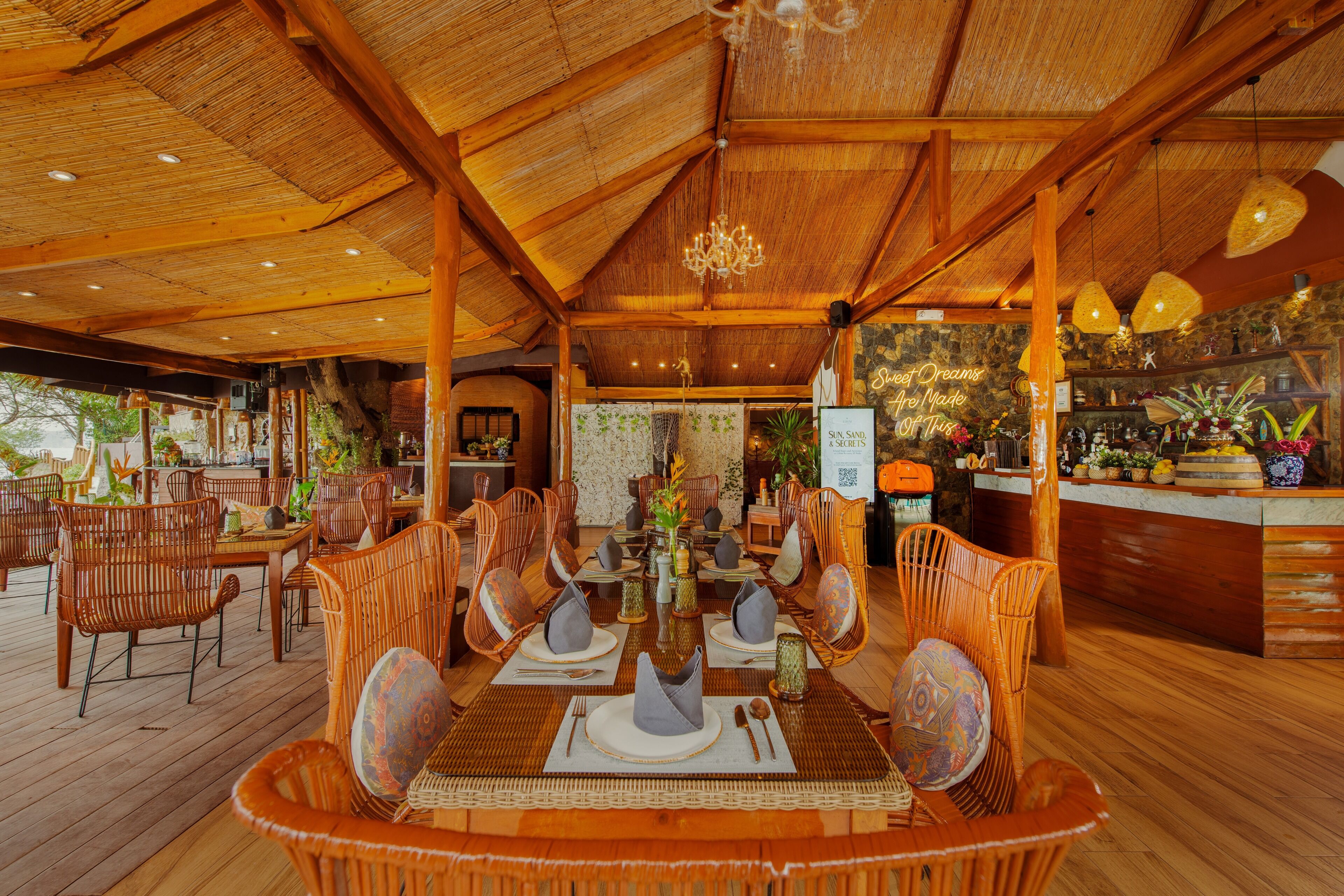 An outdoor dining area with wicker chairs and tables, a bar, and a stone wall with a sign.