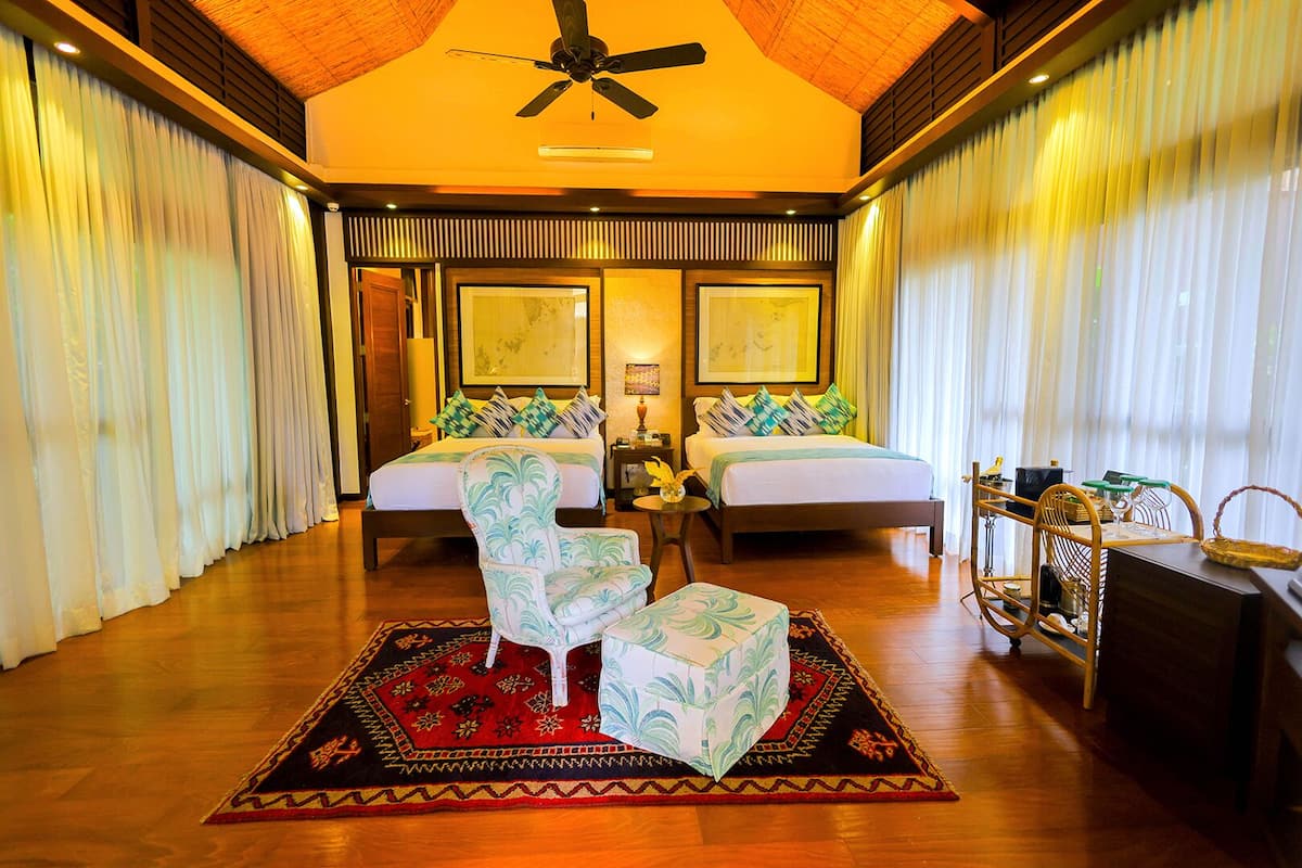 A spacious bedroom with two beds, a ceiling fan, and a large window with sheer curtains.
