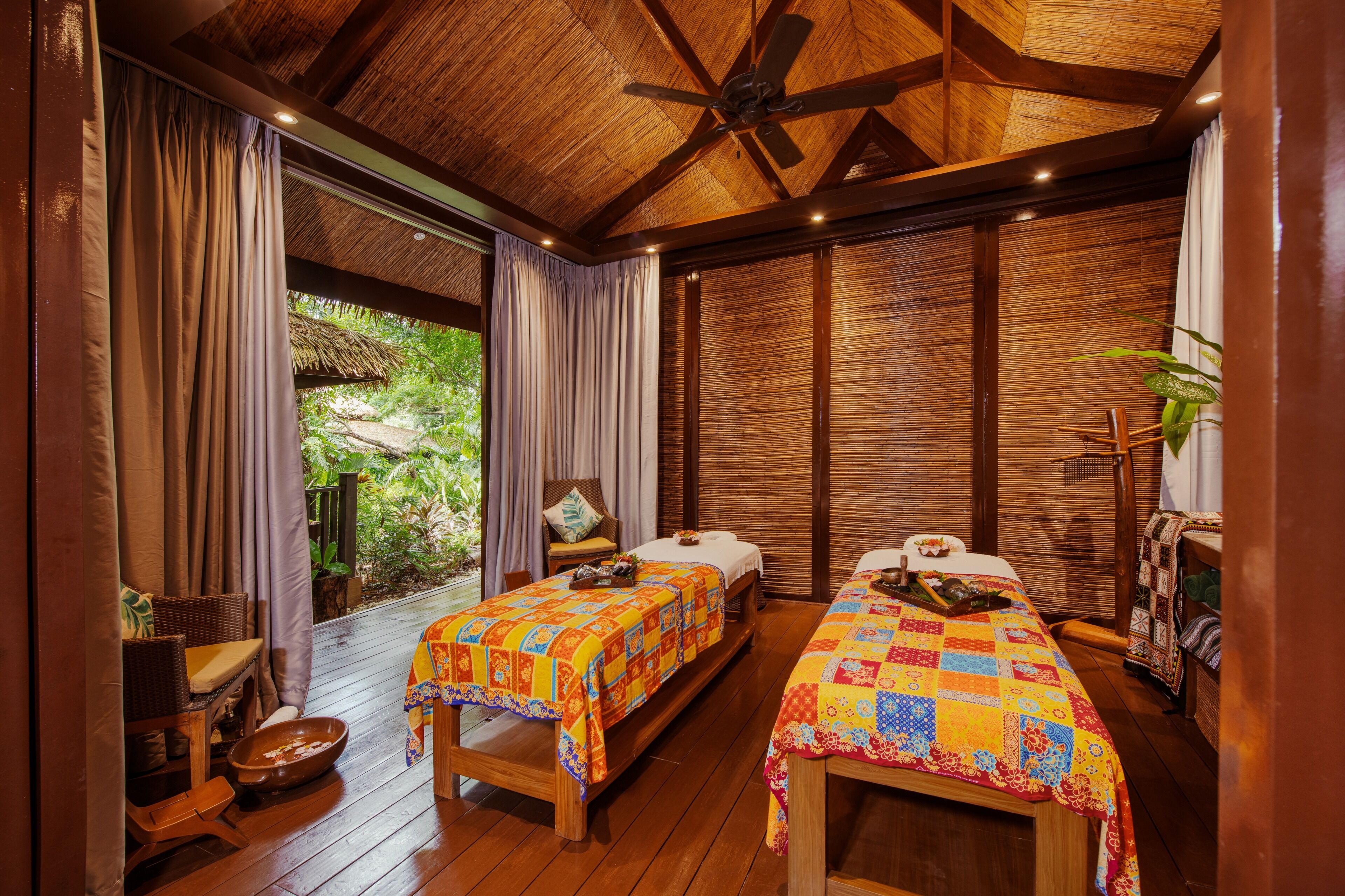 A room with two beds, wooden flooring, a ceiling fan, and a view of greenery outside.