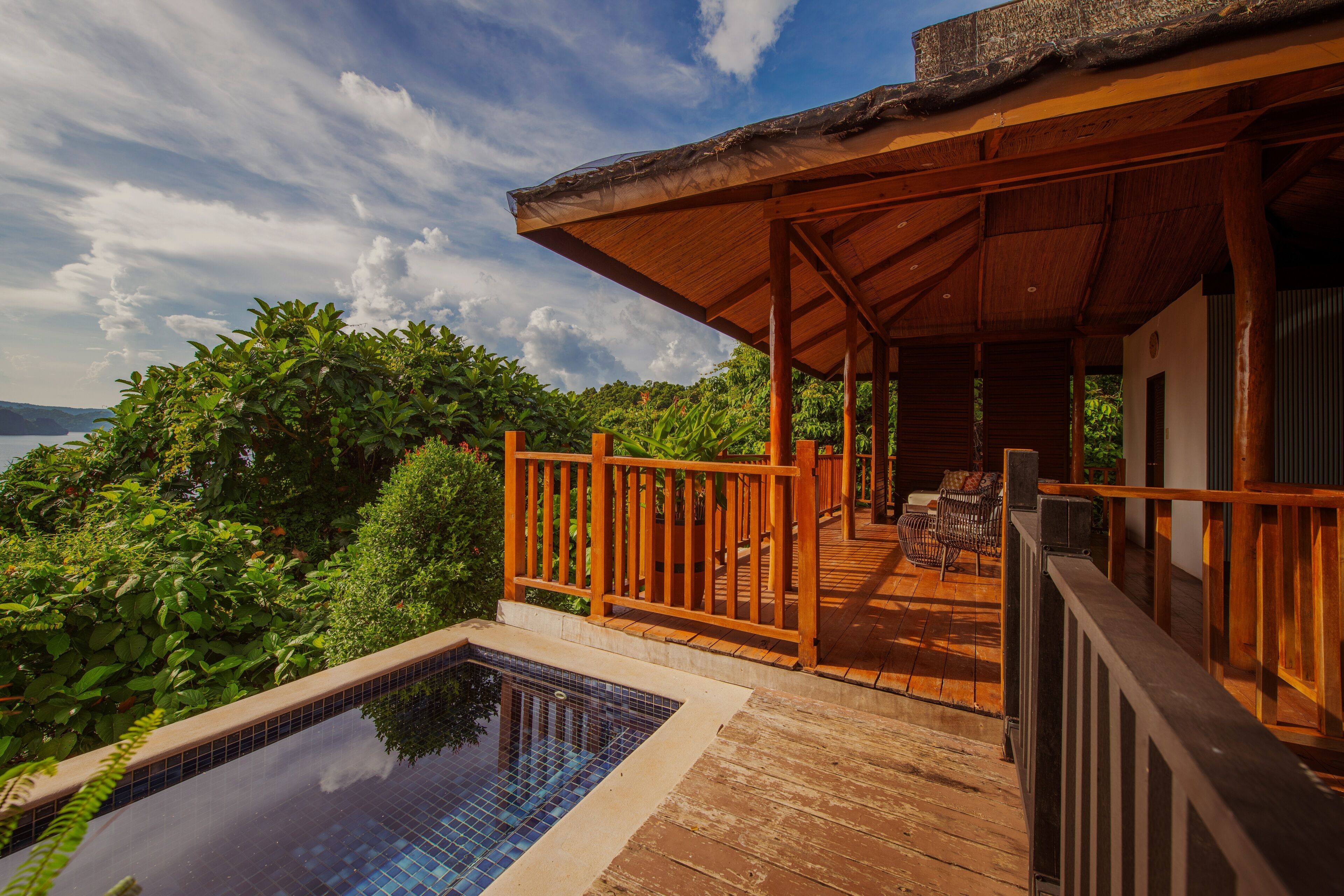 Una terraza de madera con piscina, rodeada de abundante vegetación y con vistas a un cuerpo de agua.
