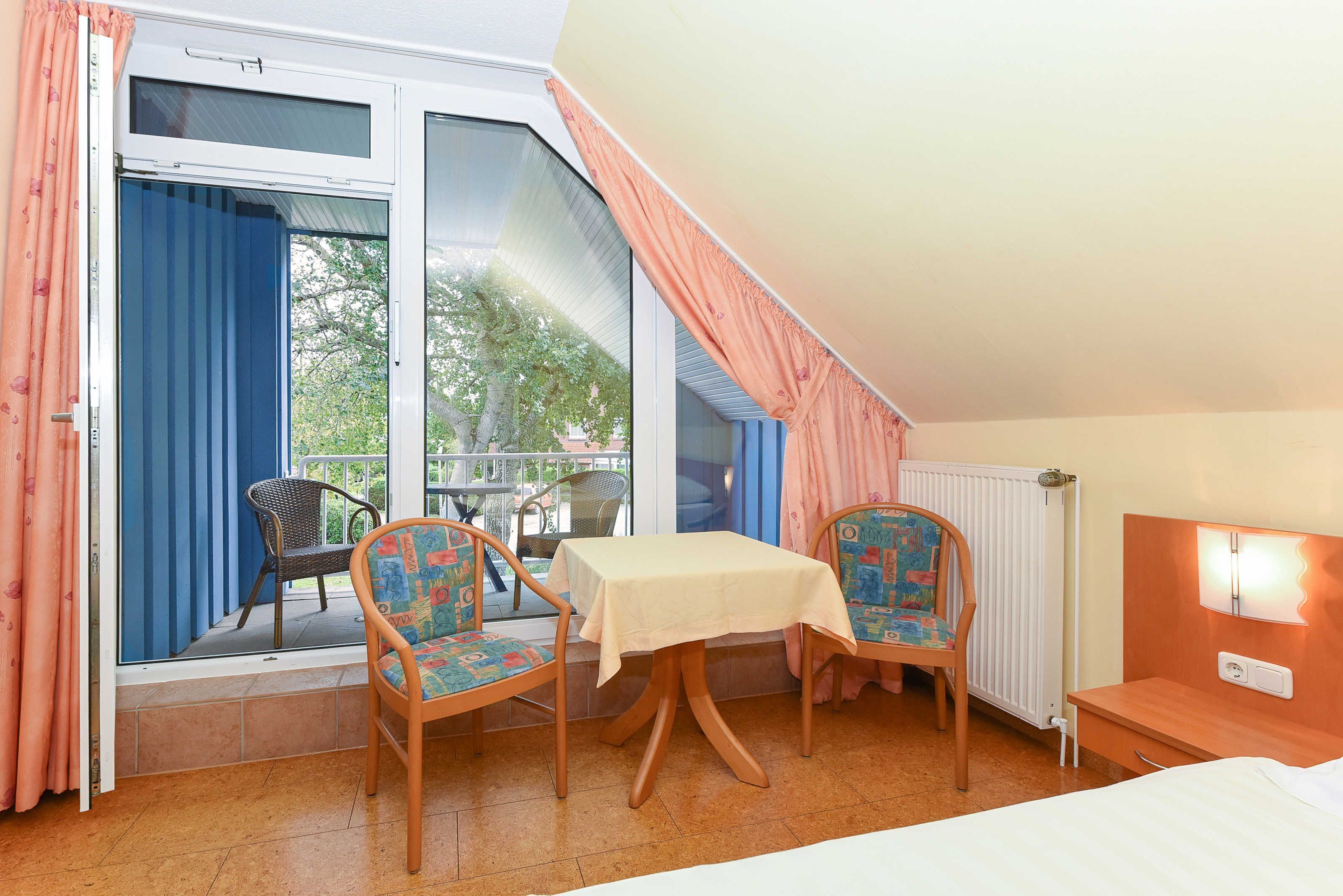 A room with a table and two chairs, a bed, a window with curtains, and a radiator.