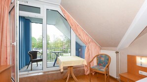 Une pièce dotée d’une porte-fenêtre coulissante donnant sur un balcon, d’une petite table recouverte d’une nappe, d’une chaise en osier, d’une table de chevet en bois et d’une table de nuit avec une lampe.