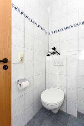 A small, modern bathroom with white tiles, a toilet, a towel rack, and a shower head.