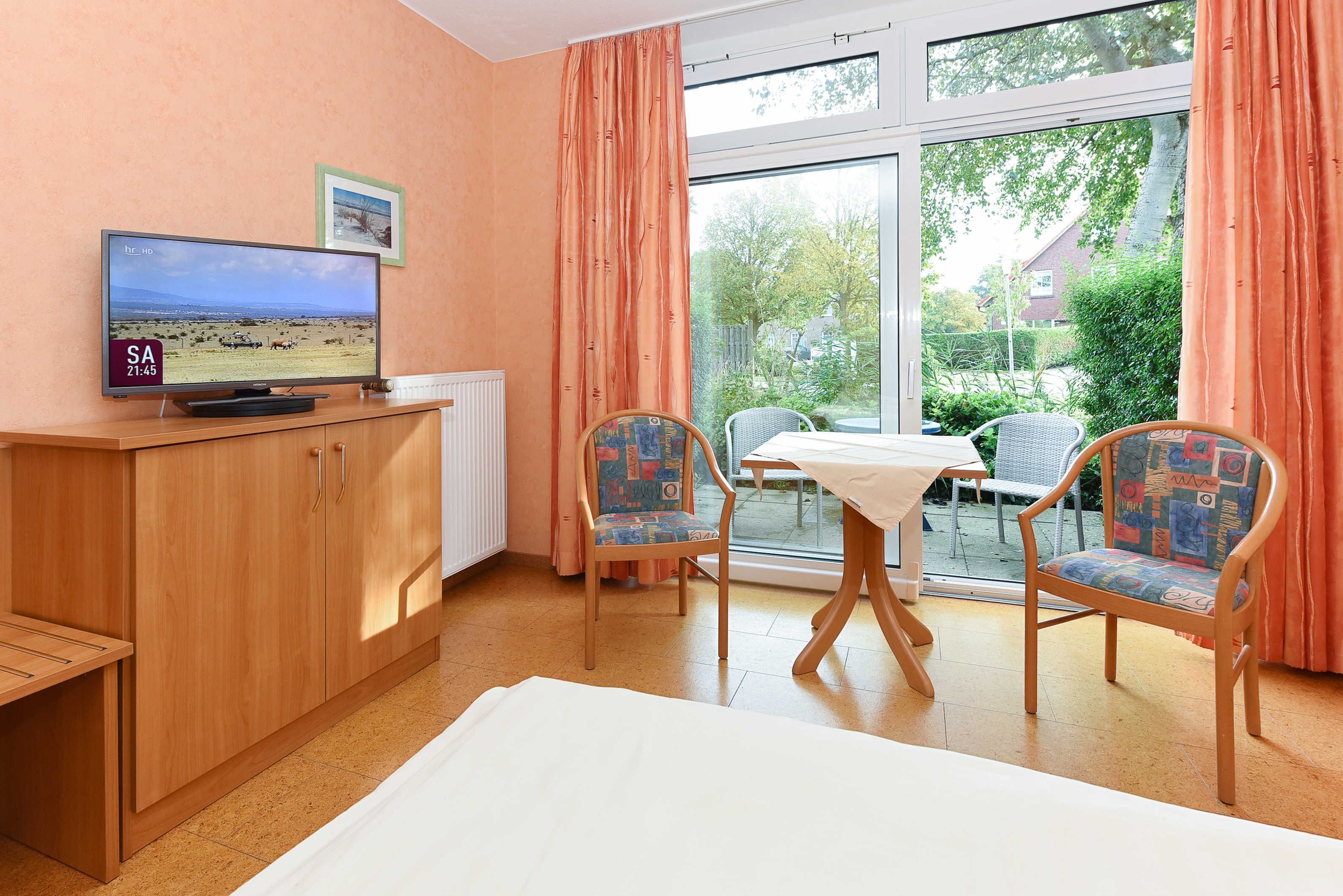 Ein Zimmer mit Fernseher, der eine Landschaft zeigt, einer Holzschrank, zwei Stühle, ein Tisch und ein Fenster mit Vorhängen.