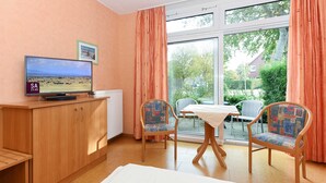 Une pièce avec un téléviseur diffusant un paysage, un meuble en bois, deux chaises, une table et une fenêtre avec des rideaux.