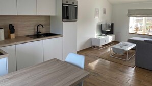 A modern kitchen with white cabinets, a black faucet, and a built-in oven. There is a dining area with a wooden table and chairs, and a living room with a gray sofa and a TV. - Kleine Ferienwohnung (Gelting)