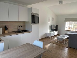 Cozinha moderna com armários brancos, uma torneira preta e um forno embutido. Há uma área de jantar com uma mesa e cadeiras de madeira, e uma sala de estar com sofá cinza e televisão.
