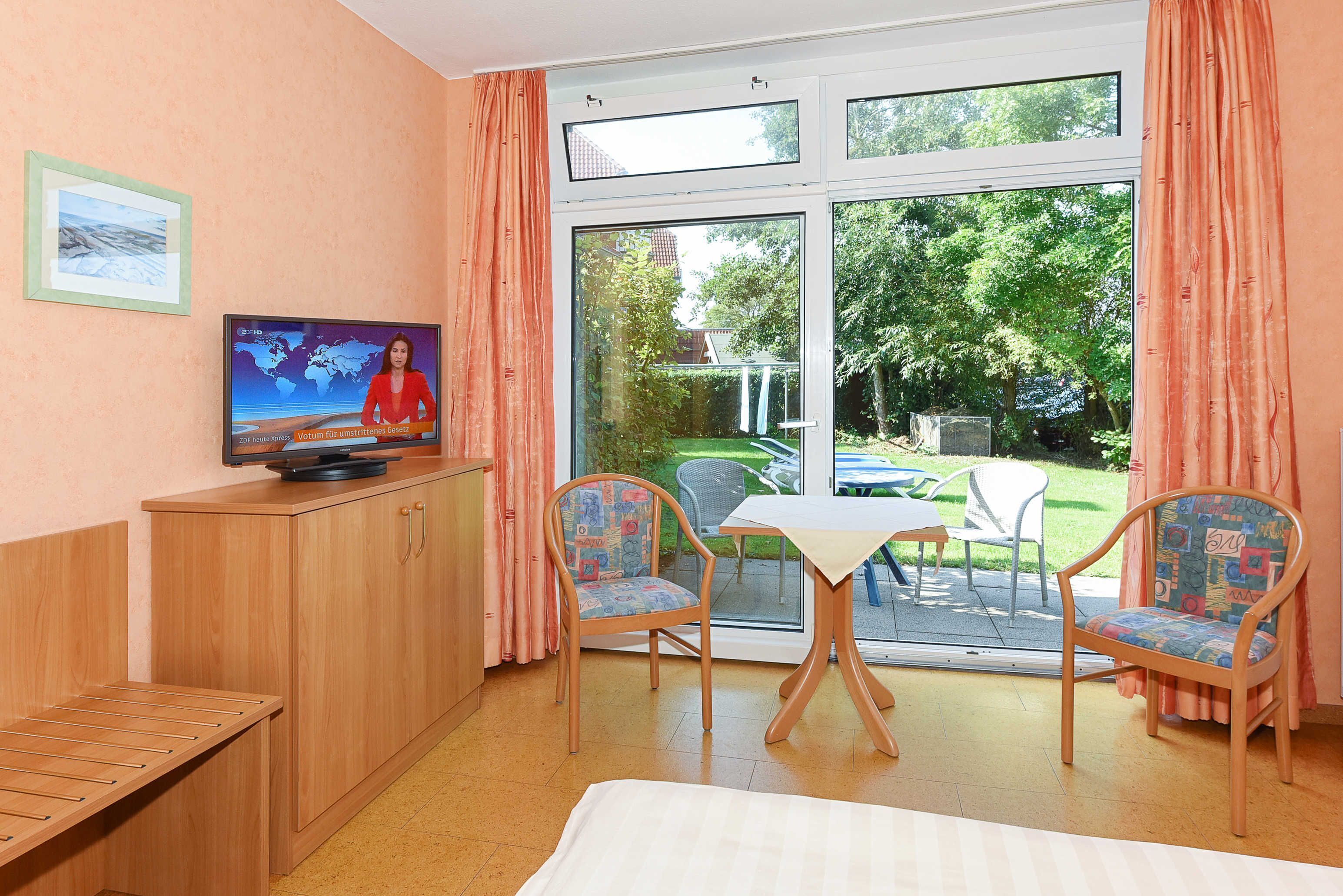 Een kamer met een bed, een televisie op een houten kast, twee stoelen, een kleine tafel en een schuifdeur naar een buitenruimte met gazon en stoelen.