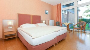 Habitación de hotel con una cama grande, una mesita de noche, una silla y una mesa junto a un ventanal amplio.