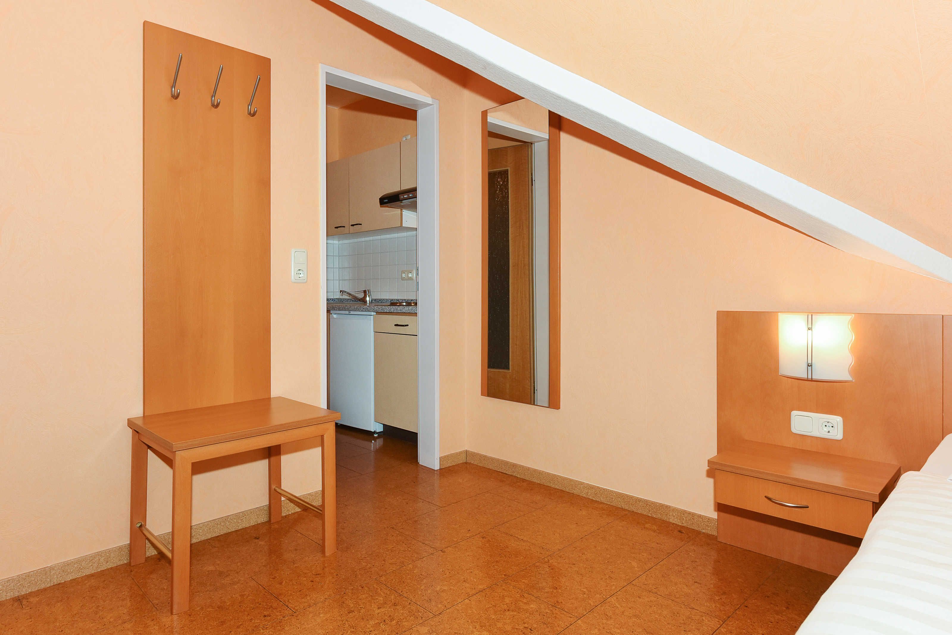 A small, well-lit room with a wooden table, a bedside table, and a mirror. There is a kitchenette visible through an open door.
