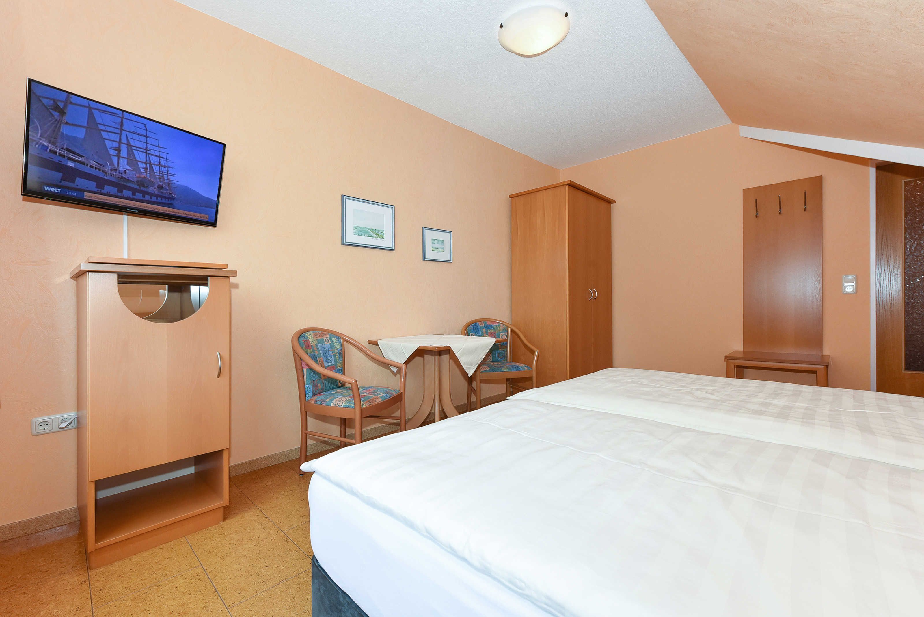 Quarto de hotel com uma cama, uma escrivaninha, duas cadeiras, uma televisão e um guarda-roupa.