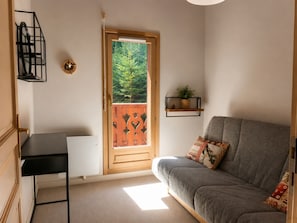 A room with a grey sofa, a small black shelf with a plant, a wooden door with a view of greenery, and a black desk.