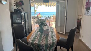 A dining area with a table set for two, a vase with flowers, and a view of the sea through an open door.