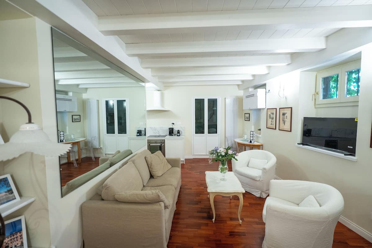 A spacious living room with a beige sofa, white armchairs, a coffee table, and a kitchen in the background.