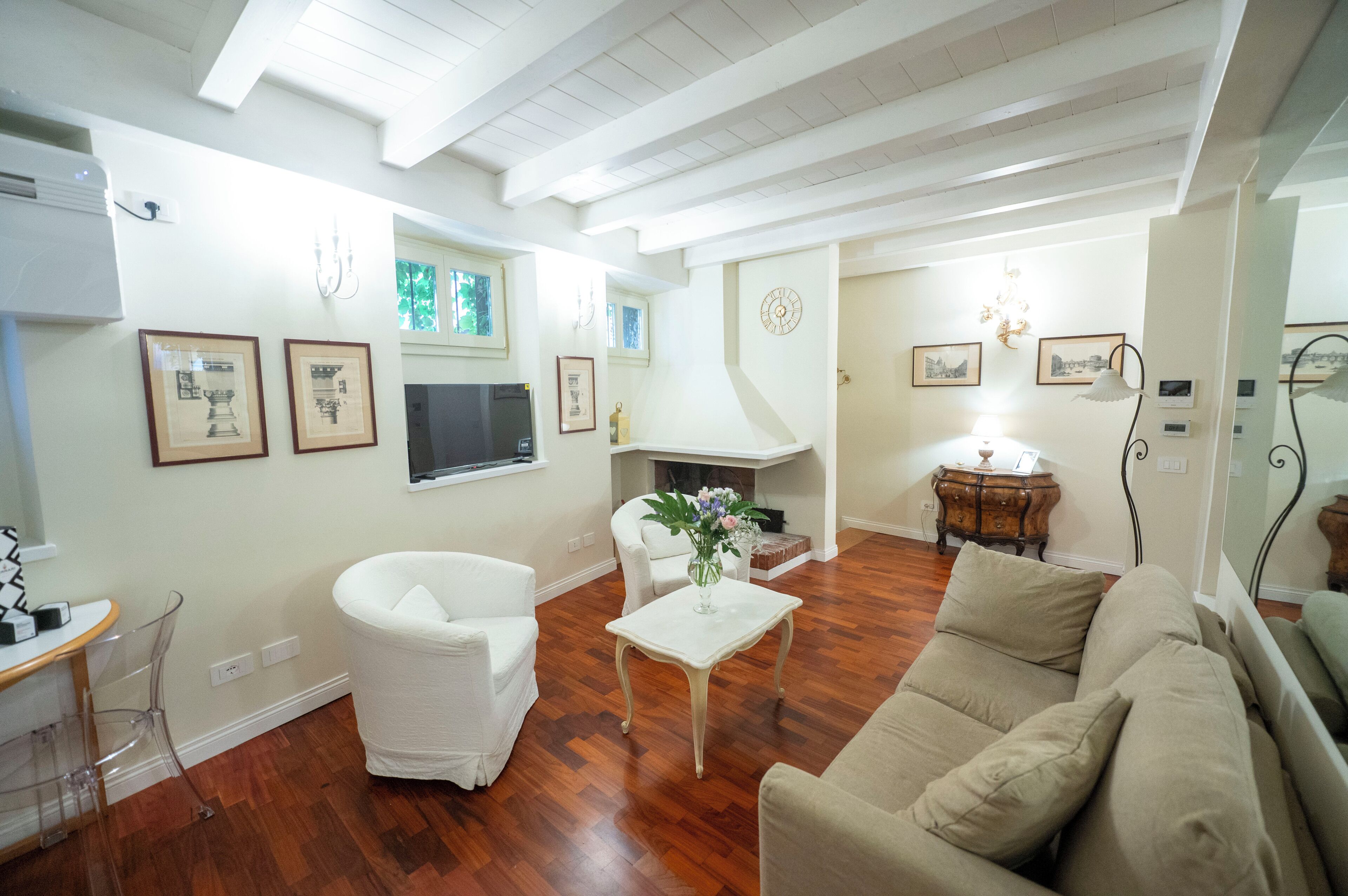 A living room with a white sofa, a beige sofa, a white armchair, a small white table, and a fireplace. There are framed pictures on the walls and a chandelier above the fireplace.