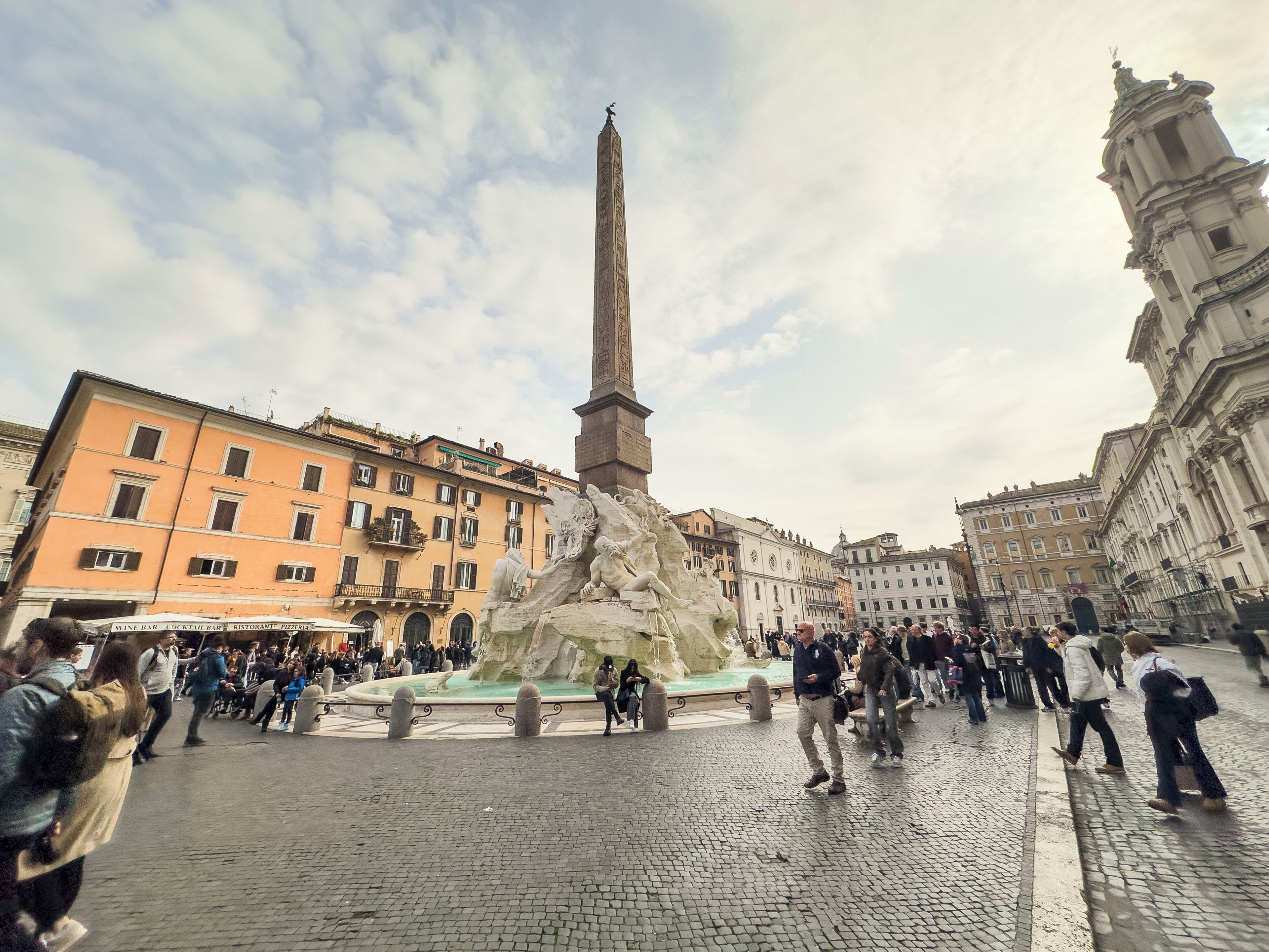 A public square with a tall obelisk monument, cobblestone pavement, and surrounding historic buildings.