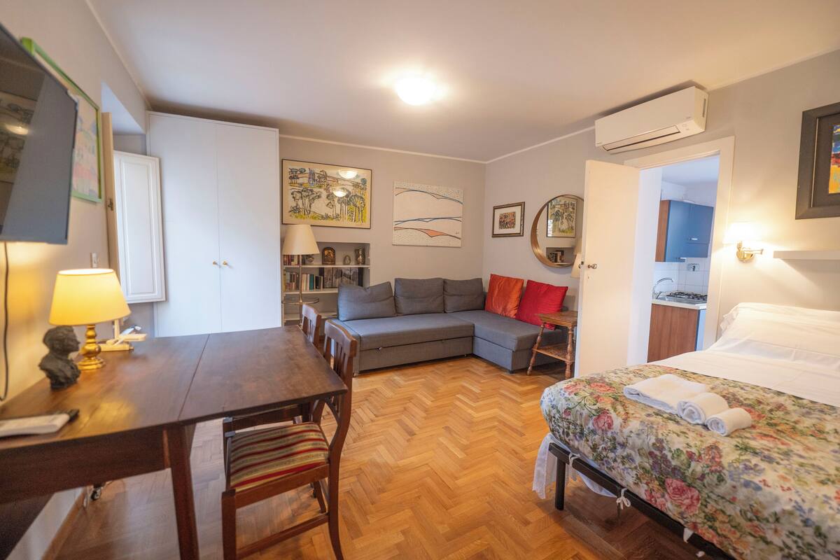 A cozy room with a wooden dining table, a sofa, a bed with floral bedding, and a small kitchen area.
