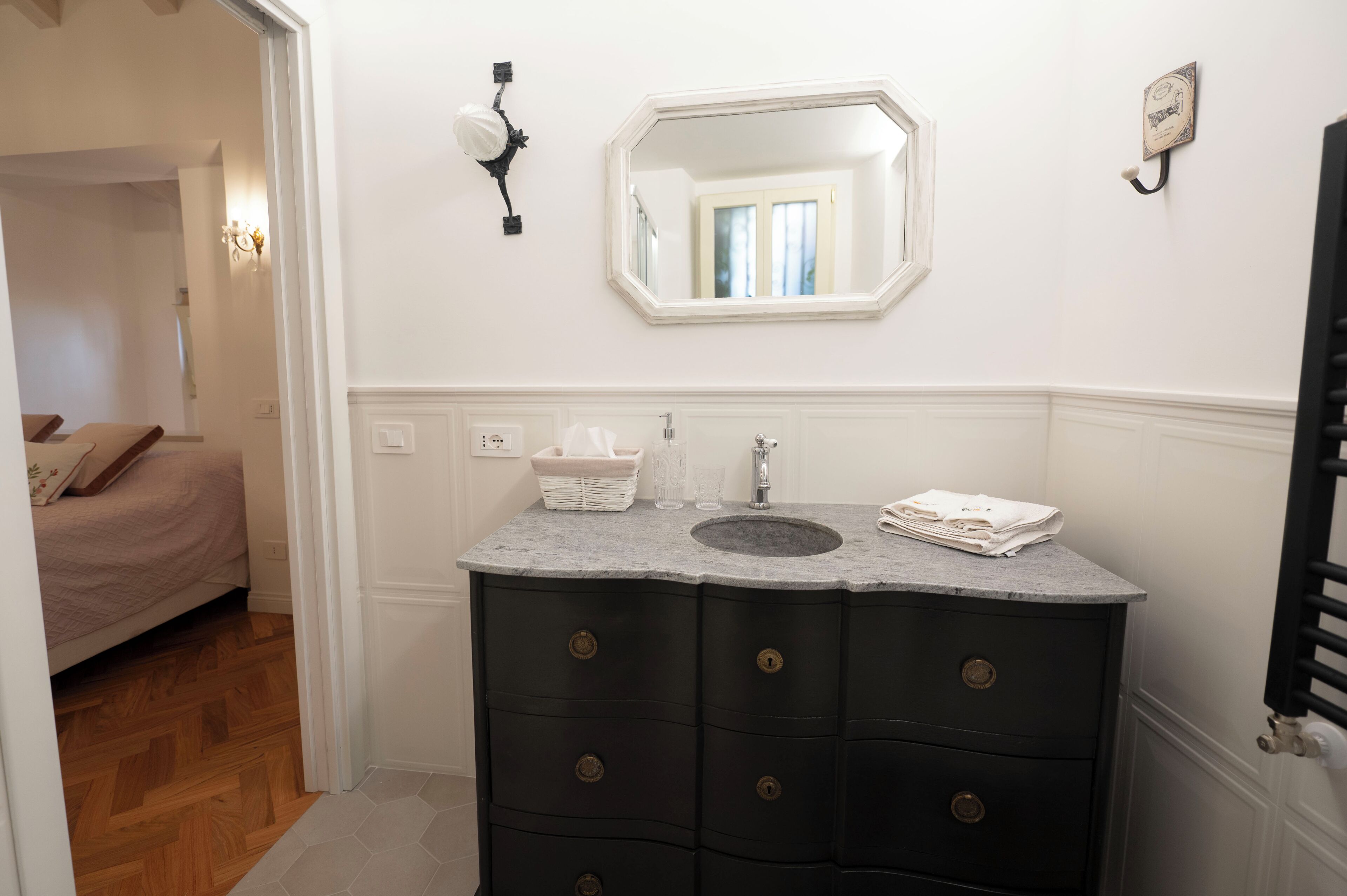 Een badkamer met een donker aanrecht, een wit wastafel en een spiegel erboven. Er is een handdoekhouder aan de muur en een raam met gordijnen.