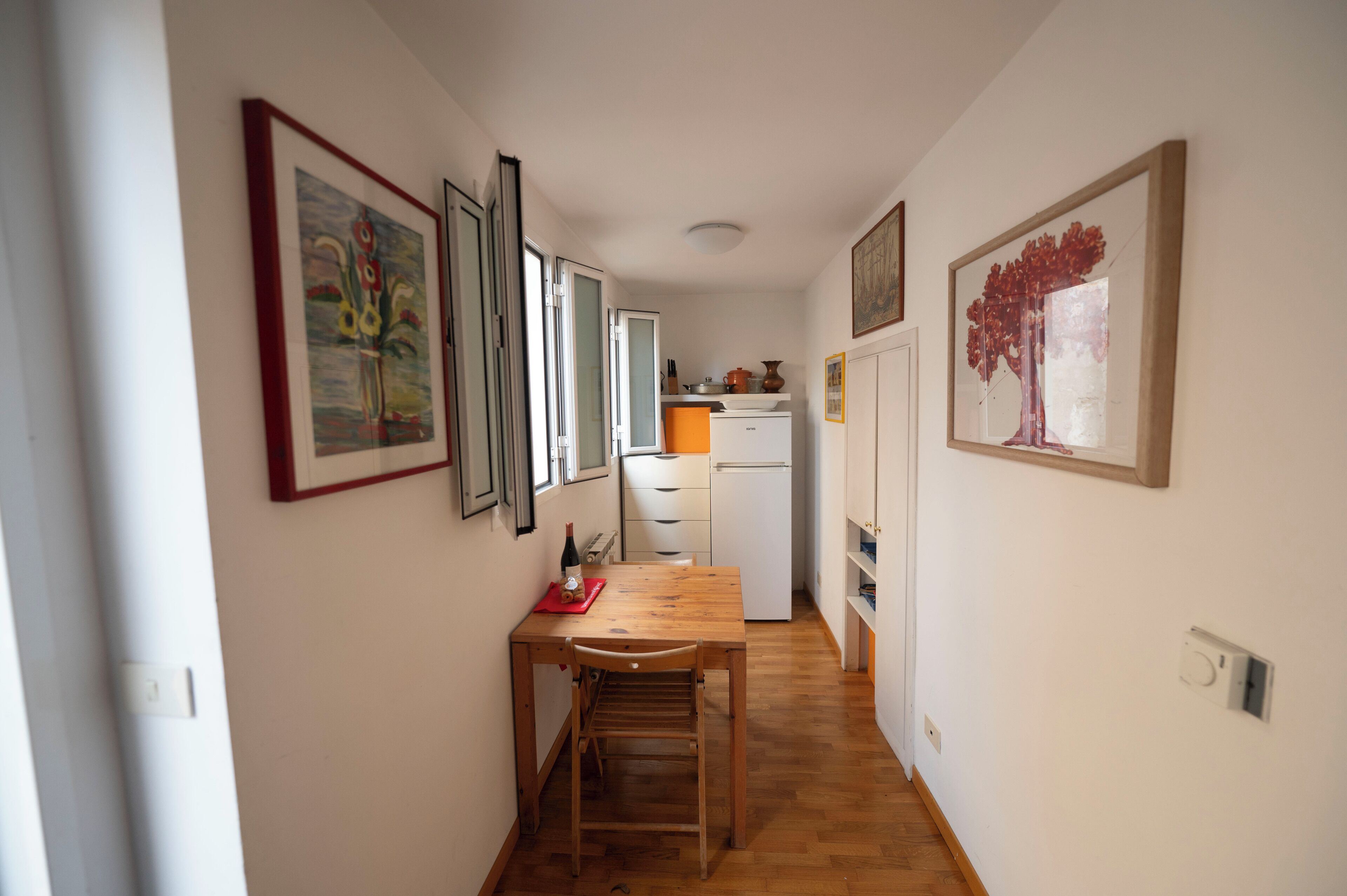 Een smalle gang met een houten tafel en stoelen, een koelkast en een deur naar een keuken.