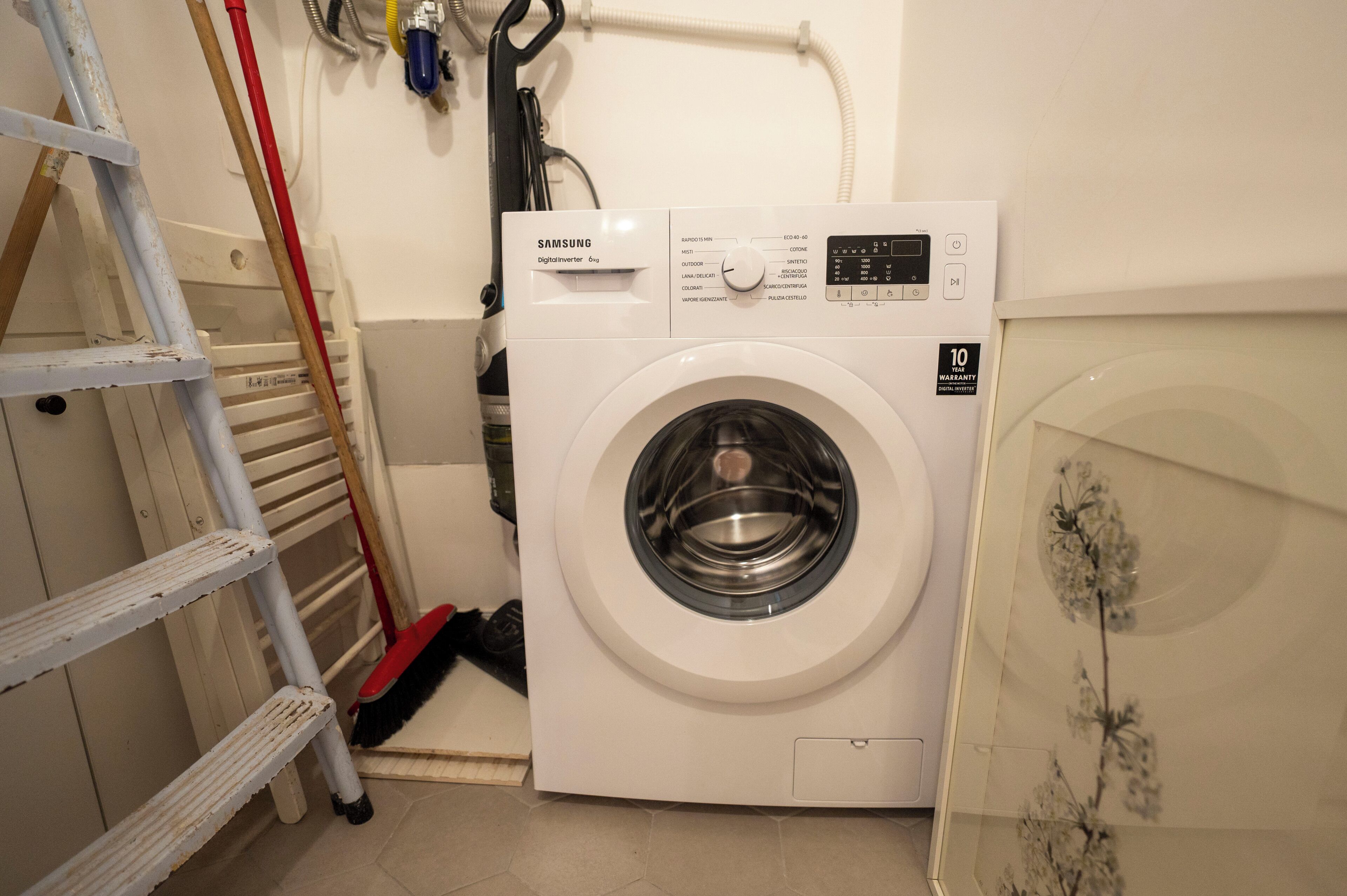 Un lave-linge Samsung compact et blanc, dans une petite buanderie équipée d’une échelle et d’outils de nettoyage.