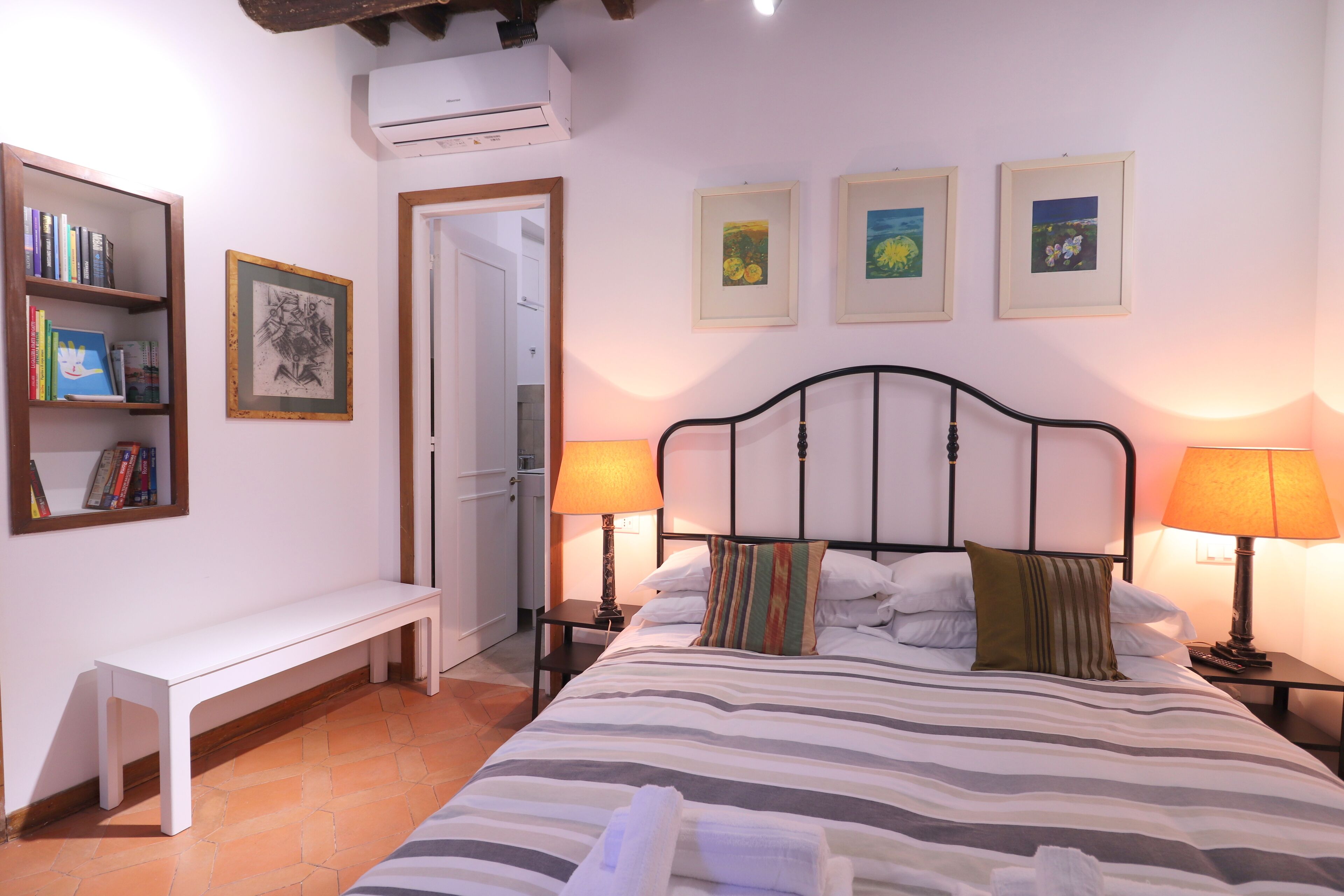 A neatly arranged bedroom with a bed, bedside lamps, a bench, a bookshelf, and framed pictures on the walls.