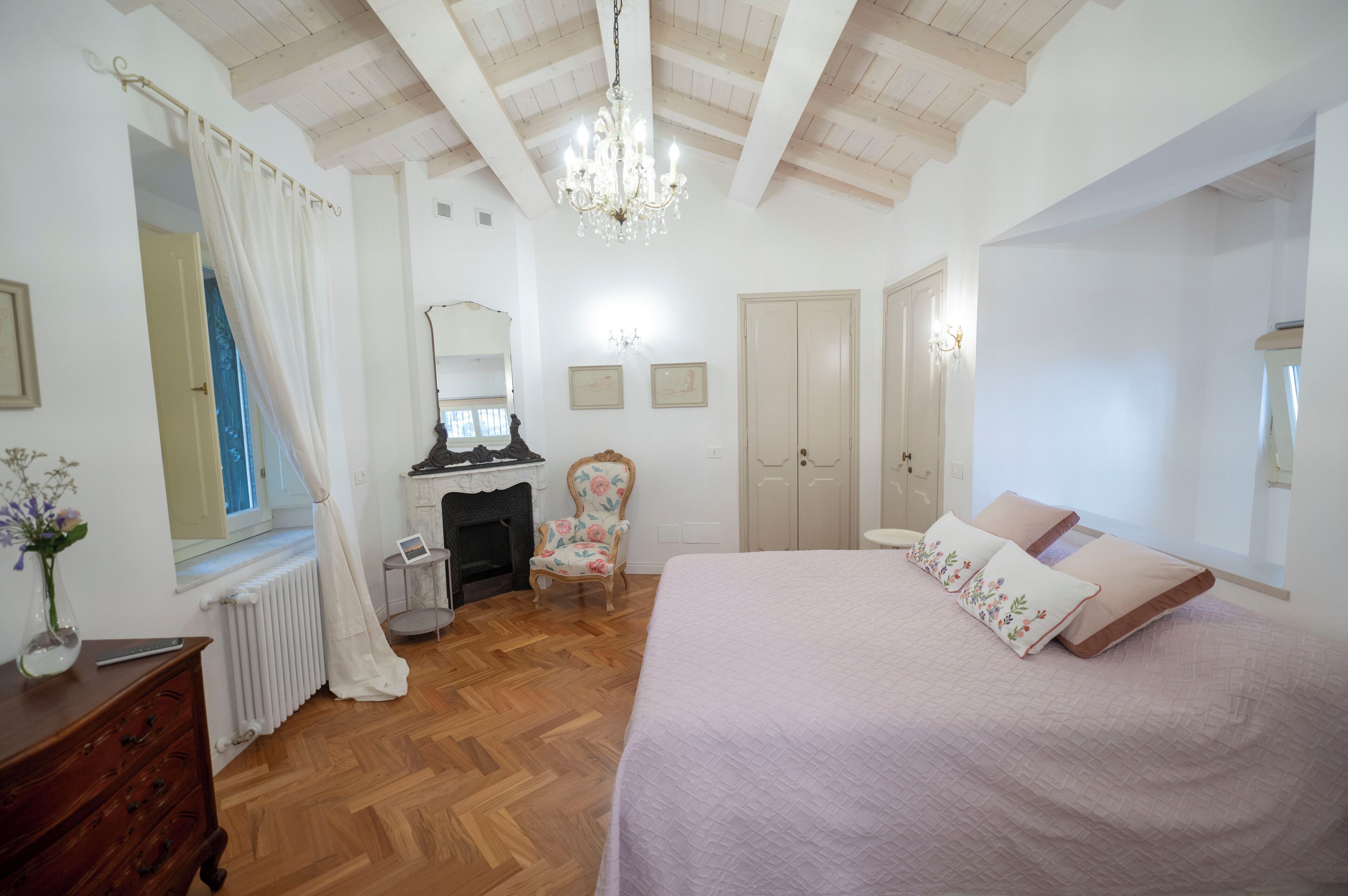 Una camera da letto con pavimento in legno, un letto con cuscini a motivi floreali, una poltrona, un camino e un lampadario.