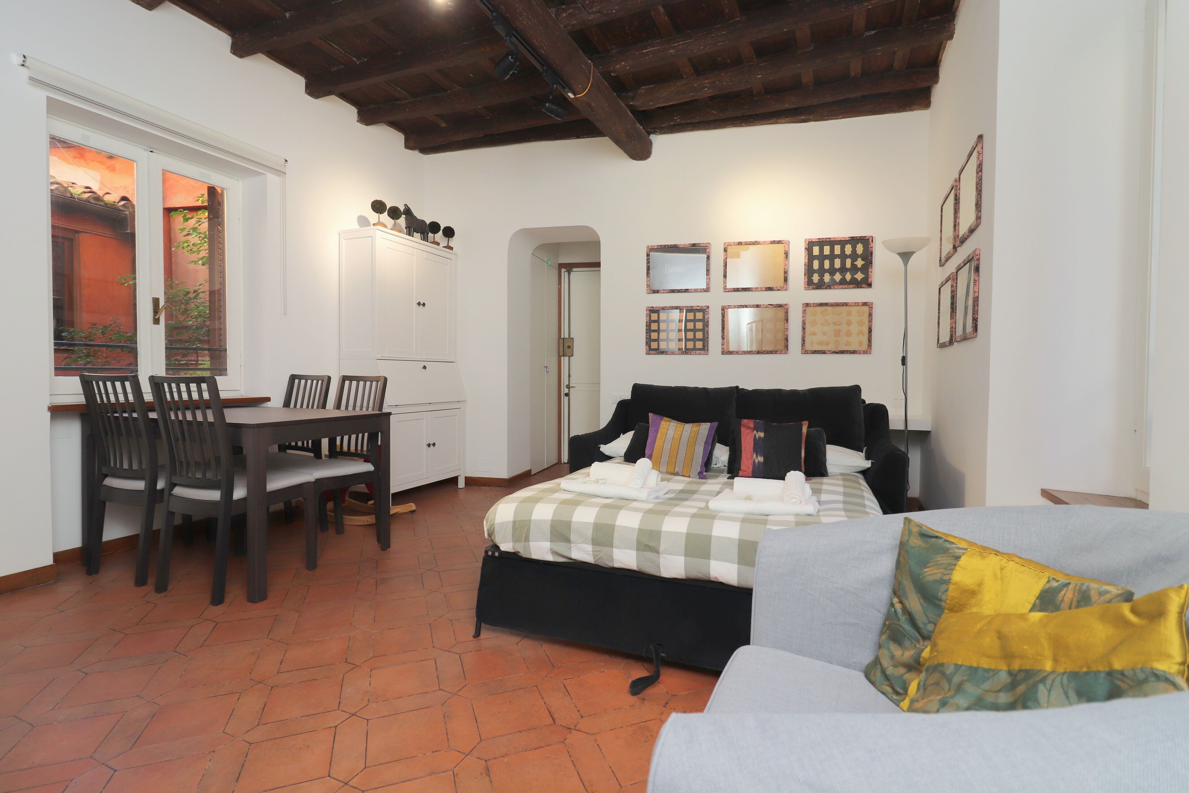 A bedroom with a bed, a sofa, a dining table with chairs, and a view of the outside through a window.