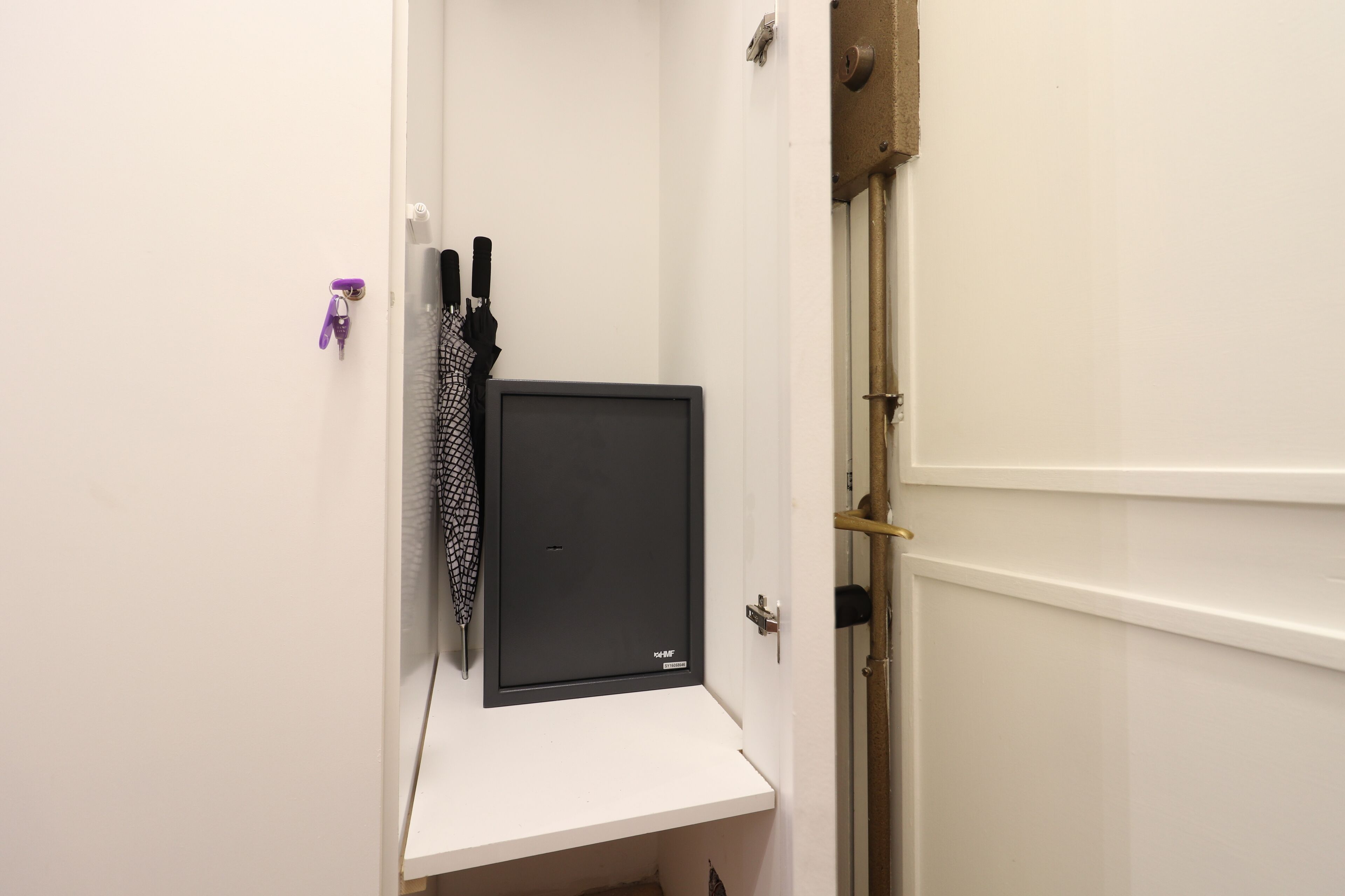 Un couloir étroit avec une porte blanche sur la droite, une petite étagère blanche sur laquelle se trouve un objet noir, et un porte-clés violet.