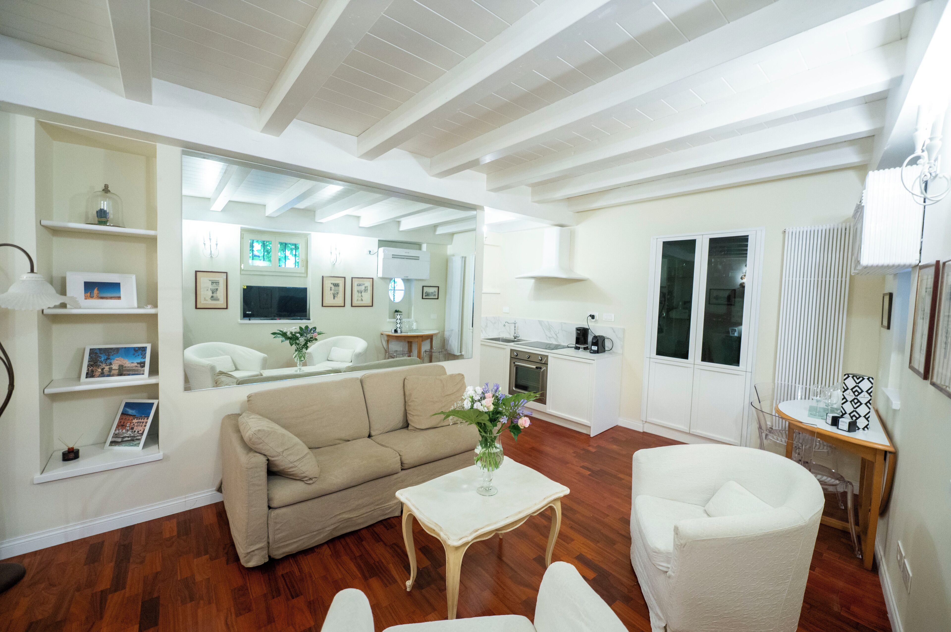 A living room with a sofa, a small table with a vase, a chair, a kitchen area, and a large mirror reflecting the room.