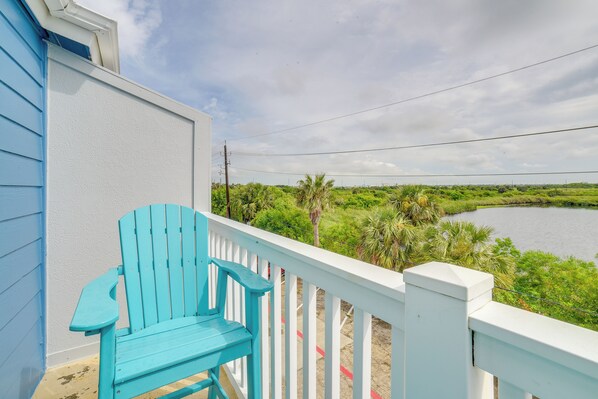 Une chaise Adirondack turquoise est posée sur la rambarde blanche d’un balcon, d’où l’on peut admirer un plan d’eau et une végétation luxuriante.