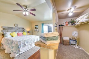 Une chambre avec un lit, un ventilateur de plafond, une table de chevet avec des serviettes, un panier en osier et un ventilateur posé sur le sol.